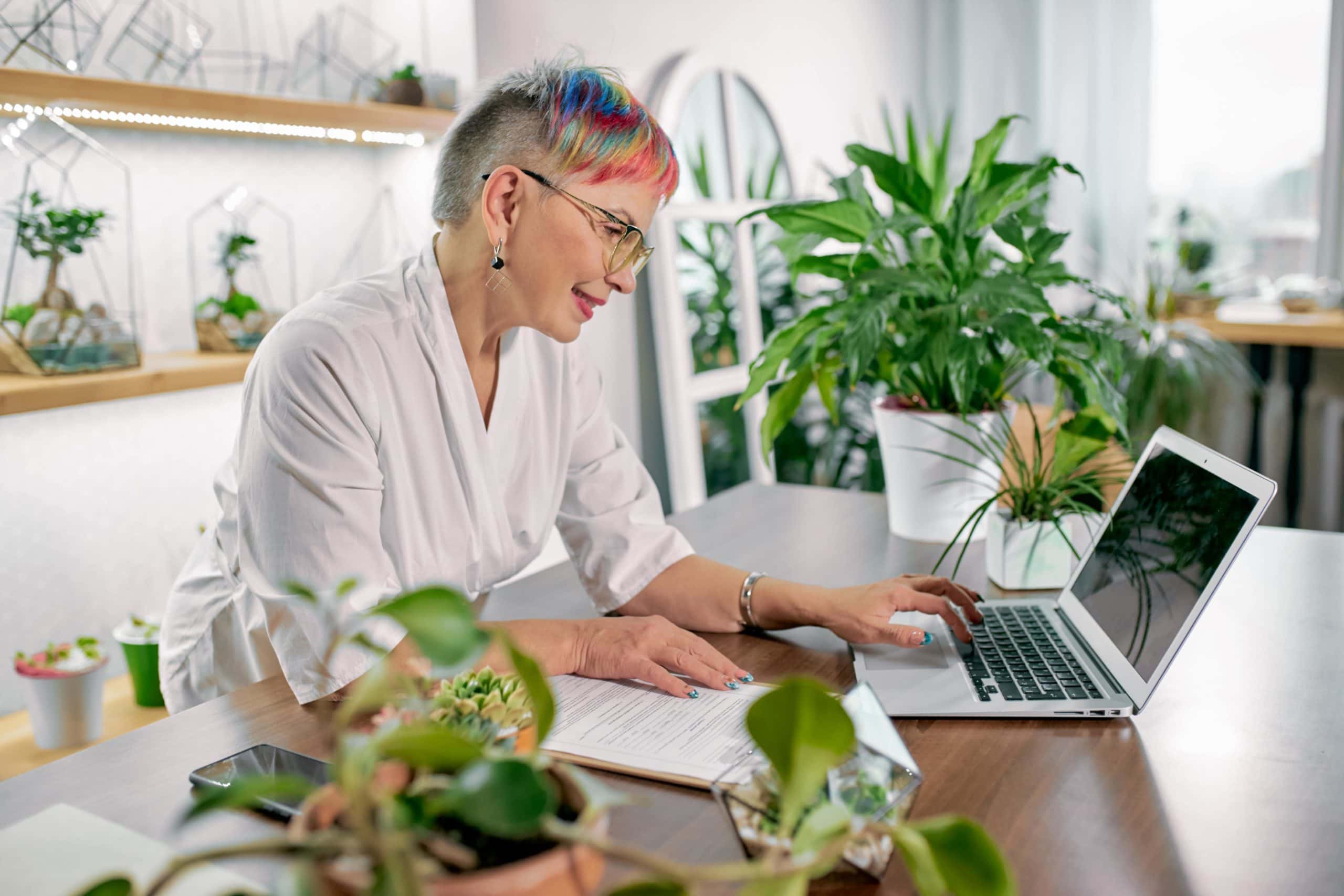 Older Woman With Colorful Hair On Laptop In Plant Shop